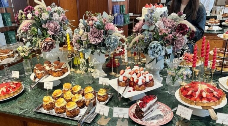 A wide 16:9 shot of a luxurious bakery display table featuring various fresh pastries, strawberry tarts, and decorated cakes, interspersed with large vintage floral arrangements and colorful spiral candles on a green marble countertop.
