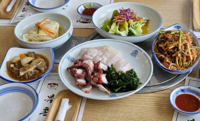 A high-angle, top-down view of a Korean dining table featuring various side dishes, including sliced raw fish, boiled octopus tentacles, seaweed, fresh green salad with purple cabbage, white kimchi, and seasoned vegetables, served in traditional blue-rimmed white ceramic bowls at Bokchon restaurant.