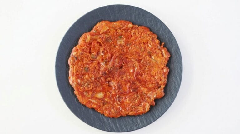 A top-down view of a round, crispy golden-brown Korean kimchi pancake served on a textured dark slate plate against a clean white background.