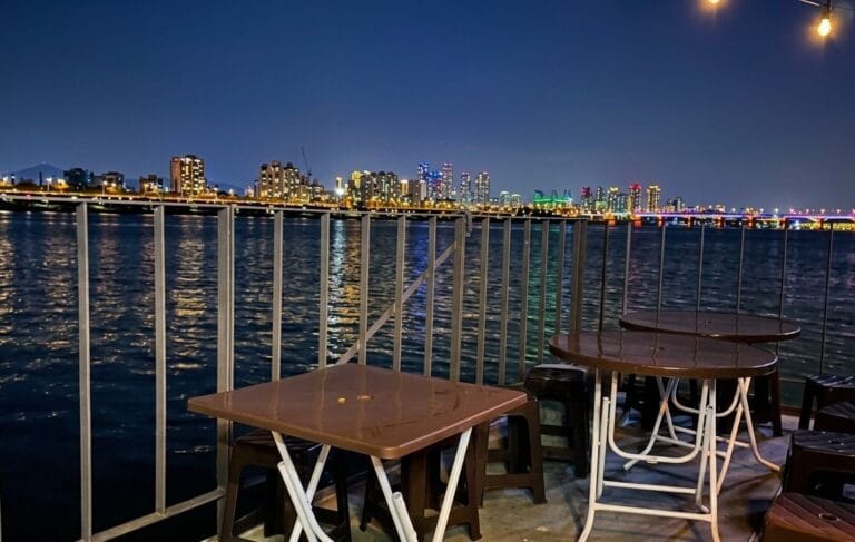 An outdoor terrace next to a river at night, featuring brown plastic folding tables and stools. A metal railing separates the terrace from the wide Han River, which reflects the colorful lights of the distant city skyline under a clear dark blue sky.