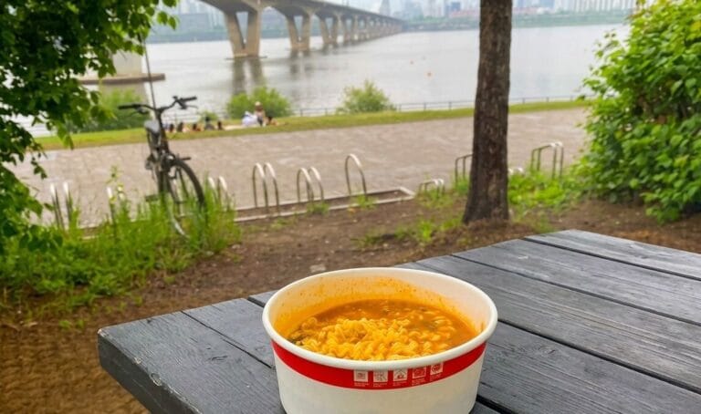 A paper bowl of instant ramen placed on a dark wooden outdoor table with a scenic view of a river and a bridge in the background.