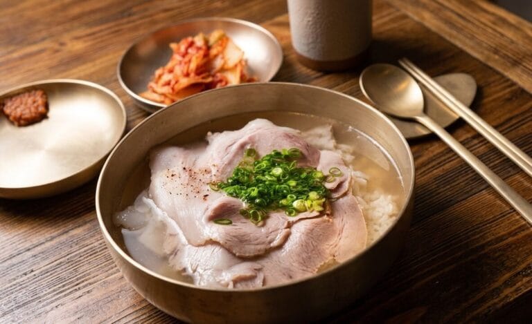 "A gourmet table setting of Dwaeji-gomtang, a clean Korean clear pork broth rice soup, with thinly sliced boiled pork and chopped green onions on top in a traditional brass bowl, served with kimchi and ssamjang on a rustic wooden table."