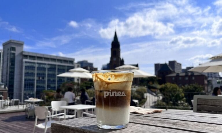 An iced layered coffee drink in a glass with 'pines' printed on it, sitting on an outdoor rooftop cafe table with a blurred city and church spire background under a blue sky.