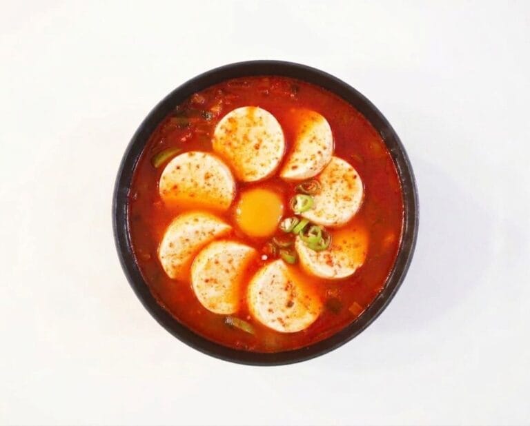 A top-down photograph of a bowl of Korean soft tofu stew (sundubu jjigae) on a white background. The stew is in a black pot with tofu arranged in a circle, a raw egg yolk in the center, and green chilies as garnish.