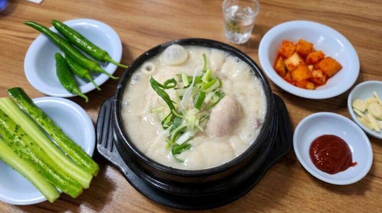 A boiling hot stone bowl of Korean perilla seed ginseng chicken soup (Samgyetang) with thick creamy white broth, topped with sliced green onions, surrounded by side dishes including fresh cucumbers, green peppers, Kkakdugi radish kimchi, garlic slices, and homemade Gochujang on a wooden table.