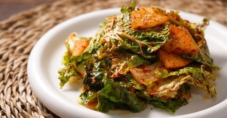 A professional close-up food photograph of Bomdong Geotjeori (Korean spring cabbage salad), featuring fresh, seasoned green leaves and sliced sweet Korean pear tossed in a vibrant red chili dressing, topped with toasted sesame seeds on a white round plate.