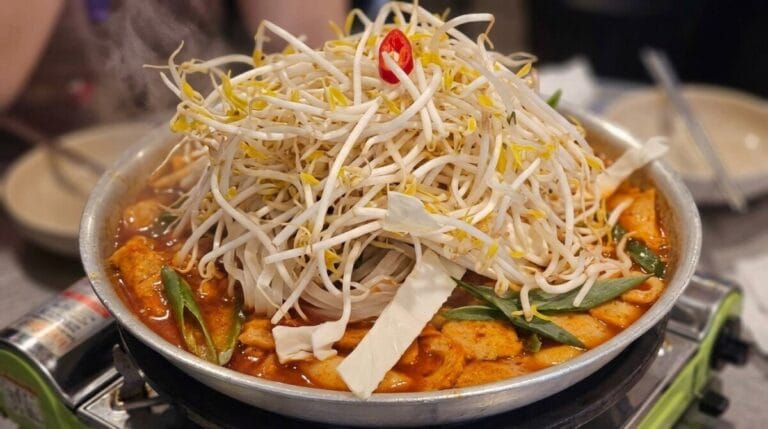 A metal hot pot of Jeukseok-Tteokbokki (Korean spicy rice cakes) simmering on a table-side gas stove, piled high with fresh green perilla leaves, cabbage, and a boiled egg in a bubbling red chili sauce.