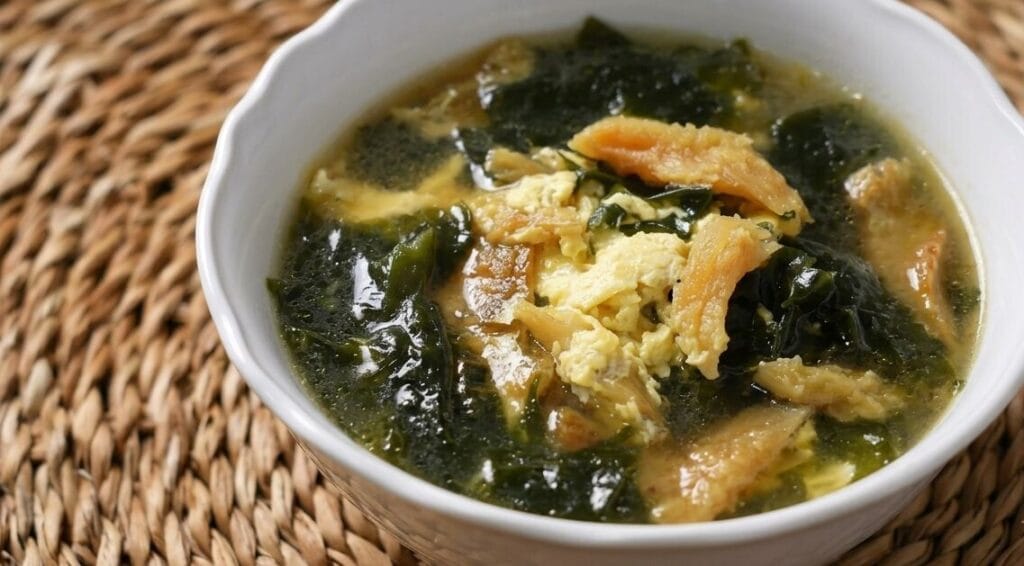 A close-up, high-angle shot of a white ceramic bowl filled with Korean seaweed soup (miyeok-guk) containing scrambled eggs and dried pollack, placed on a woven rattan placemat.
