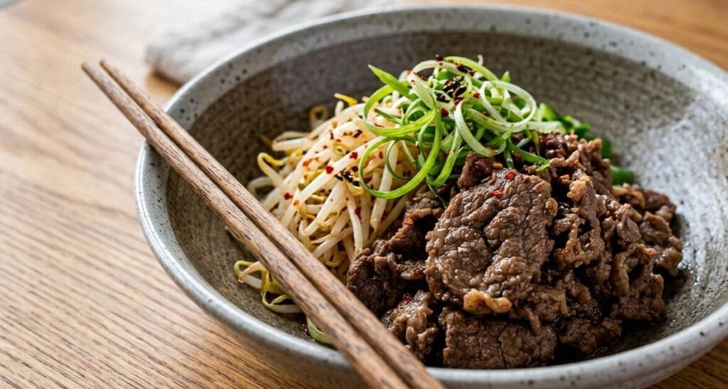 A close-up, high-angle shot of a Korean bulgogi dish served in a speckled grey ceramic bowl on a light wooden table. The bowl contains seasoned beef bulgogi on the right, blanched bean sprouts (sukju-namul) on the left, and is topped with finely shredded green onions and red pepper flakes. A pair of wooden chopsticks rests across the edge of the bowl, with soft natural light illuminating the scene from the left.