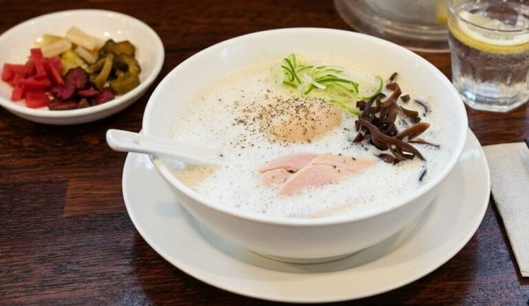 A high-angle, close-up shot of a bowl of creamy Japanese ramen served on a dark wooden table. The ramen is topped with a soft-boiled egg, sliced pork, wood ear mushrooms, and thinly sliced scallions. Next to the bowl is a small side dish of pickled vegetables and a glass of water with a lemon slice. insadong