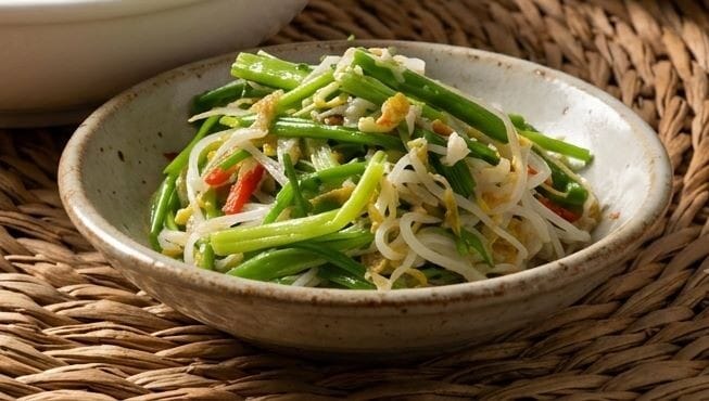 A professional food photograph featuring Korean Sukju Minari Muchim (seasoned mung bean sprouts and water dropwort). A large white ceramic bowl filled with the vibrant green and white seasoned vegetables is topped with thinly sliced red chili peppers. In the foreground, a smaller plate holds a separate portion of the dish, all styled on a textured woven straw mat over a dark wooden table under soft, cinematic lighting.