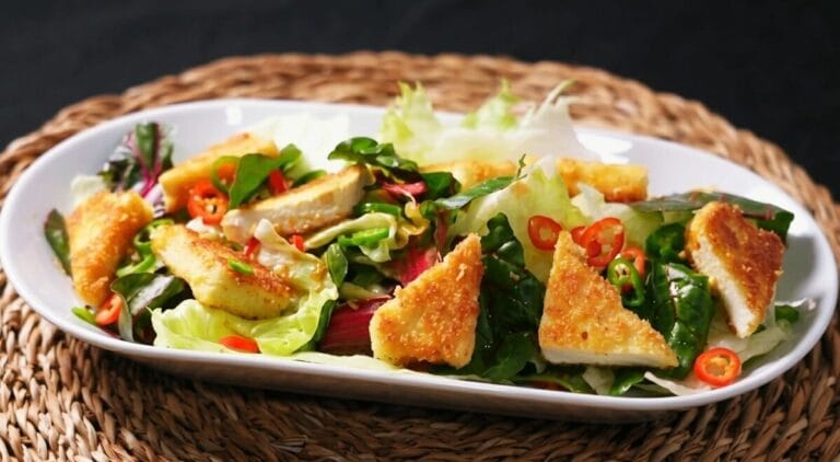 A professional close-up photograph of crispy fried tofu salad (Tofu Yuringi), featuring golden panko-crusted tofu cubes on a bed of fresh mixed greens and red chard, garnished with red and green chili peppers on a white plate.