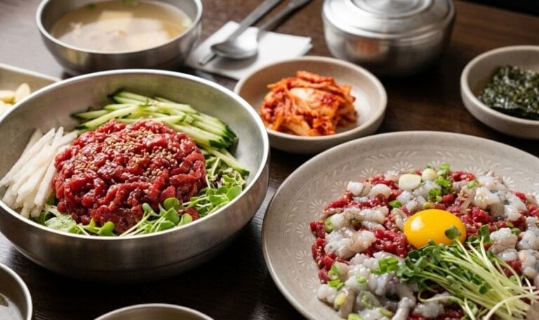 A close-up shot of a Korean dining table featuring two main dishes from Buchon Yukhoe in Gwangjang Market: a large metal bowl of Yukhoe-bibimbap (Korean beef tartare rice bowl) topped with sliced cucumber and pear, and a ceramic plate of Yukhoe-tangtangi (beef tartare mixed with live octopus) topped with a raw egg yolk and sprouts. Traditional side dishes like kimchi and soup are visible in the background.