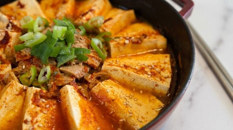 A tight, close-up photograph of Korean tuna and tofu stew (Jjageuli) in a dark red pot. Thick slices of firm tofu are neatly arranged around a savory center of seasoned canned tuna and kimchi, all simmering in a rich, spicy red broth. The dish is garnished with a mound of freshly chopped green onions and a sprinkle of sesame seeds. The pot rests on a clean, modern white marble countertop.