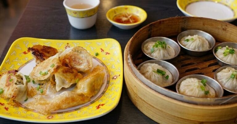 Two styles of Korean-Chinese dumplings from Gubok Mandu served on a table: pan-fried dumplings with a crispy skirt on a yellow plate and steamed dumplings (Xiao Long Bao) in a bamboo steamer.