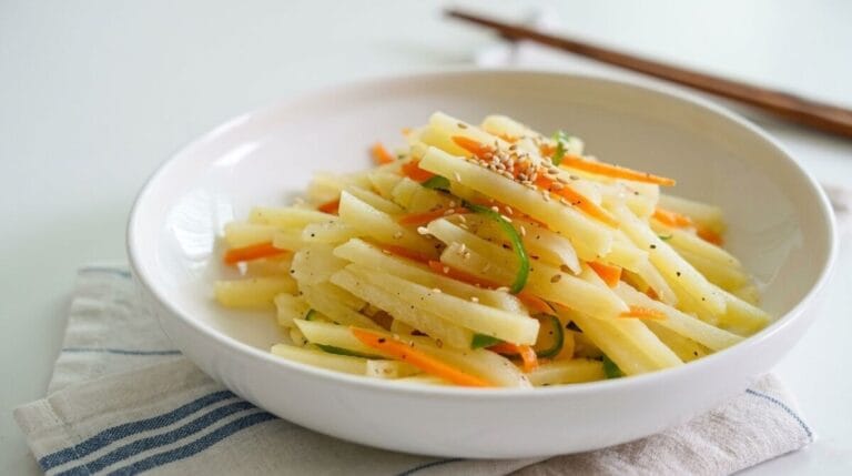 A close-up shot of Gamja Bokkeum (Korean stir-fried potato side dish) with julienned potatoes, carrots, and green chilis in a white ceramic bowl, garnished with sesame seeds on a white table.